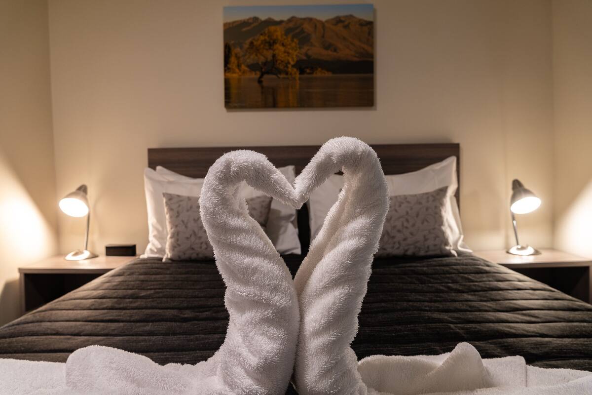 A bed with a dark quilt and two white towels arranged in a heart shape. There are two bedside lamps and a painting on the wall.