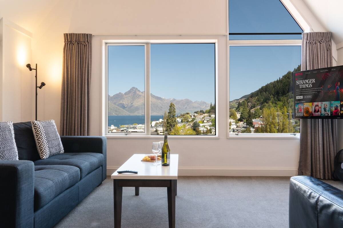 A modern living room with a sofa, a coffee table with a bottle and glasses, and a large window with a mountain view.