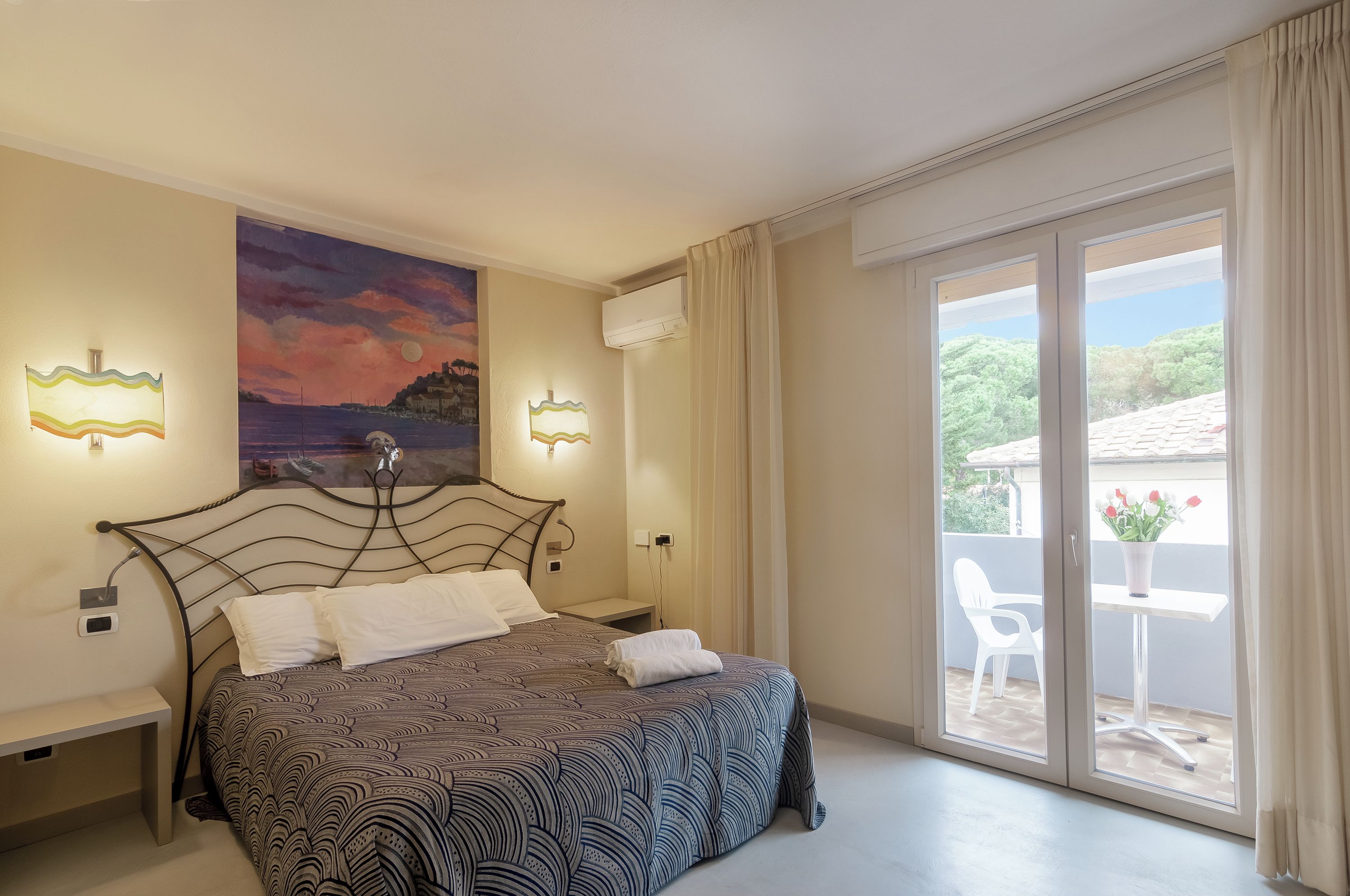 A bedroom with a bed, a painting, two wall-mounted lamps, a window with curtains, and a view of the outside.