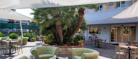 An outdoor seating area with cushioned chairs and tables, surrounded by potted plants and palm trees.