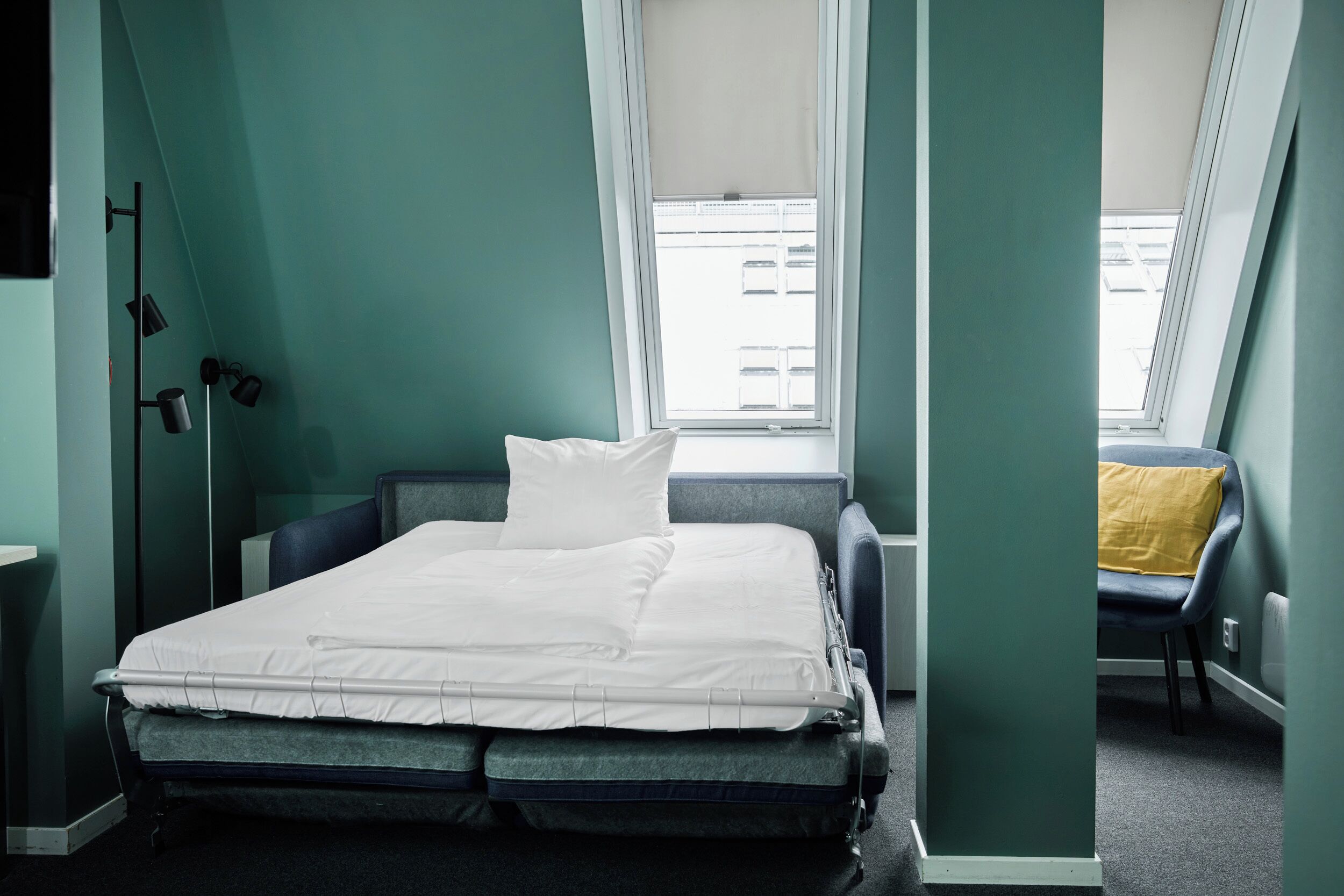 A hospital room with a bed, a window with a view of buildings, and a chair with a yellow cushion.