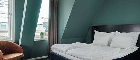 Una cama bien hecha en una habitación con paredes azul verdoso, una ventana con cortinas y una silla.