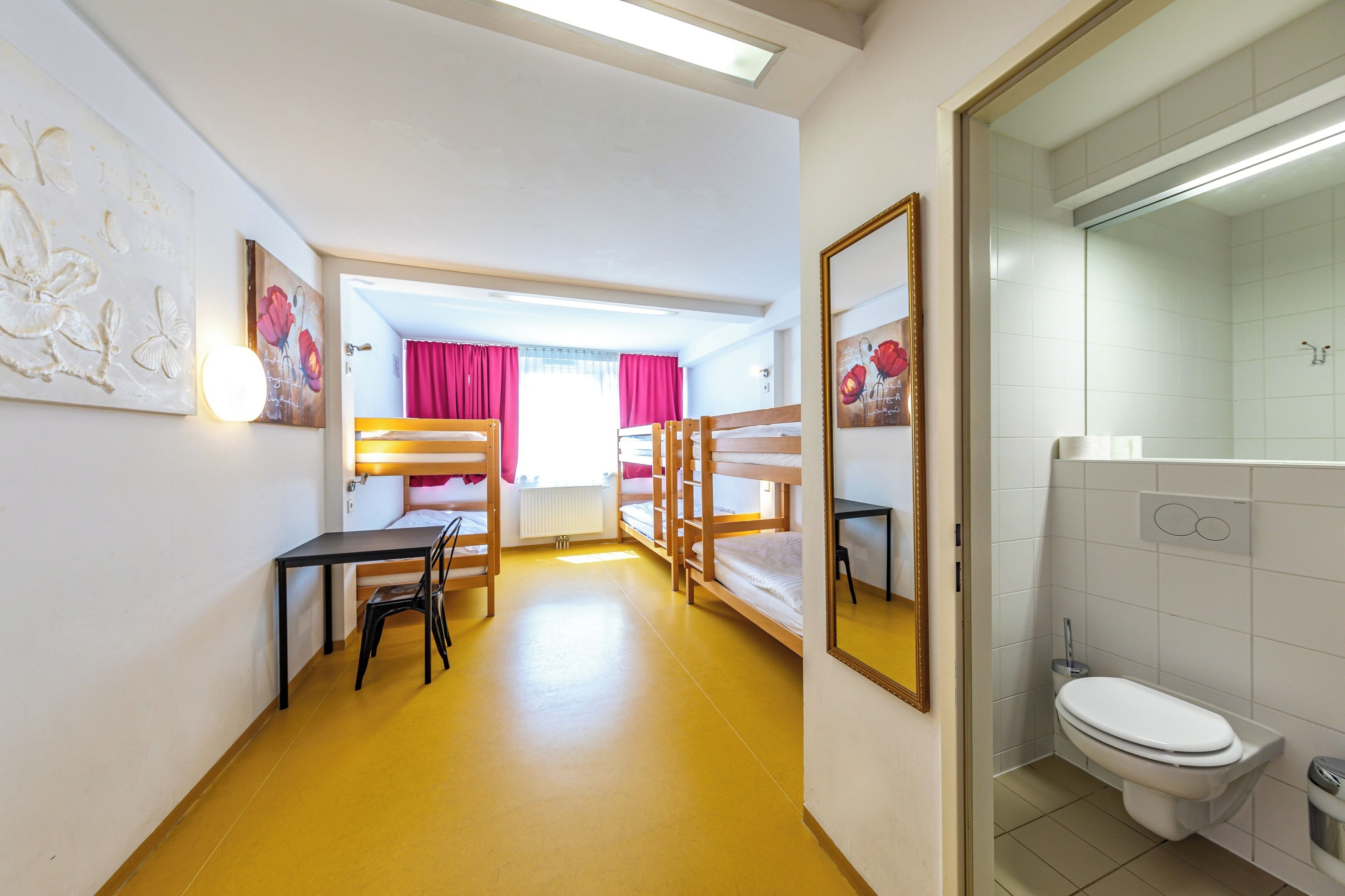 A room with bunk beds, a desk, chairs, a mirror, and a bathroom with a toilet and sink.