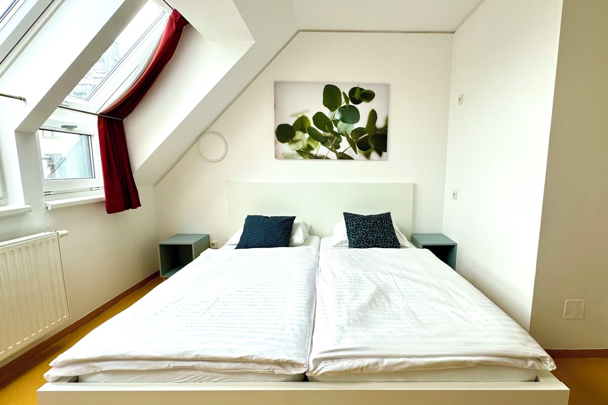 A bedroom with a large bed, two bedside tables, a window with red curtains, and a wall-mounted plant.