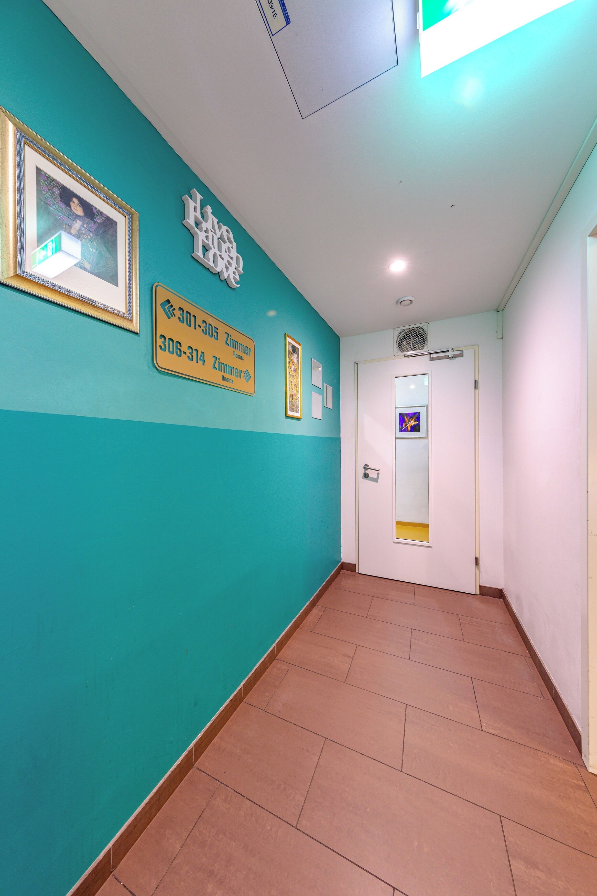 Un pasillo con paredes azul verdosas, suelo de baldosas y una puerta con un espejo. Hay carteles con números de teléfono y un cuadro en la pared.
