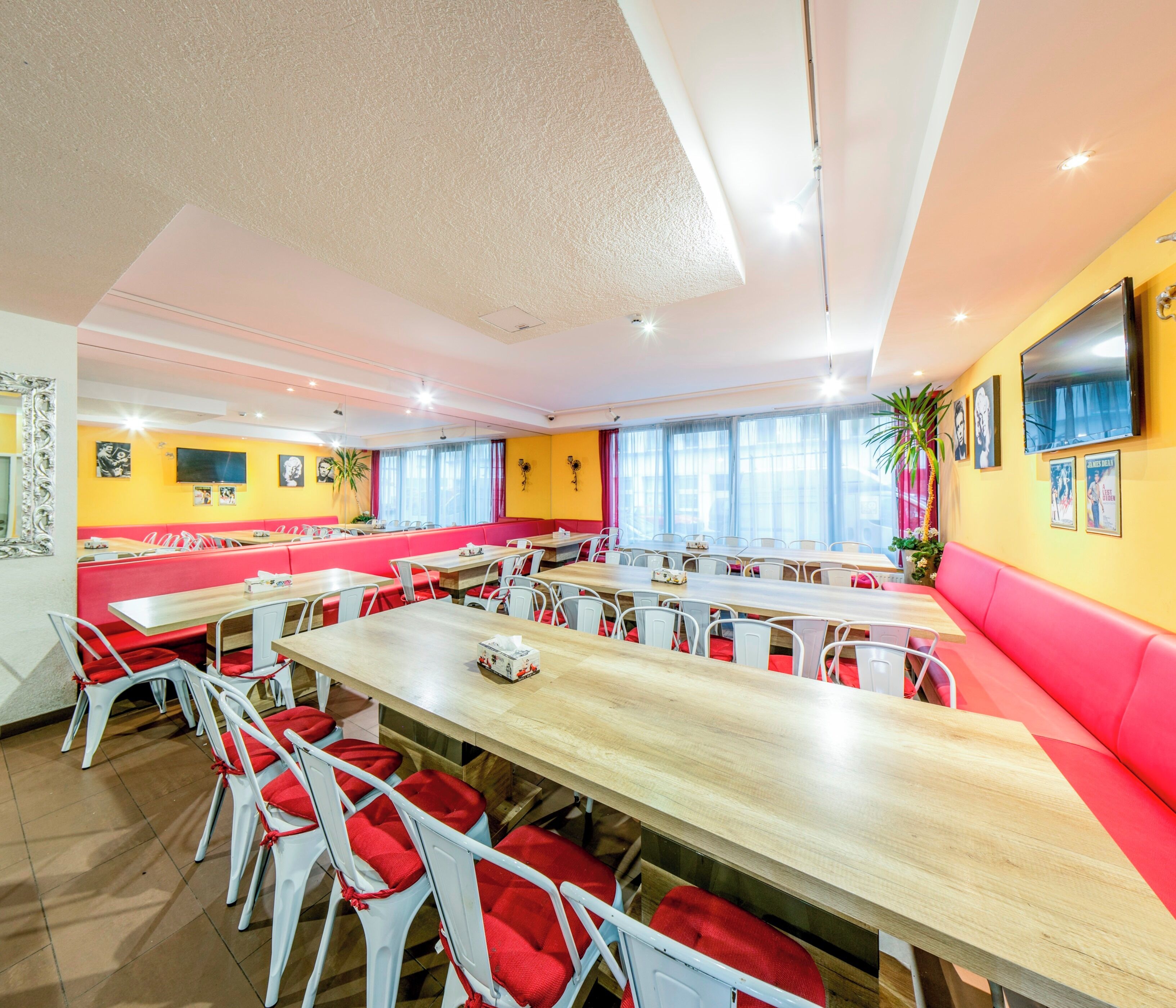 A spacious dining area with multiple tables and chairs, a large window with curtains, and a television mounted on the wall.