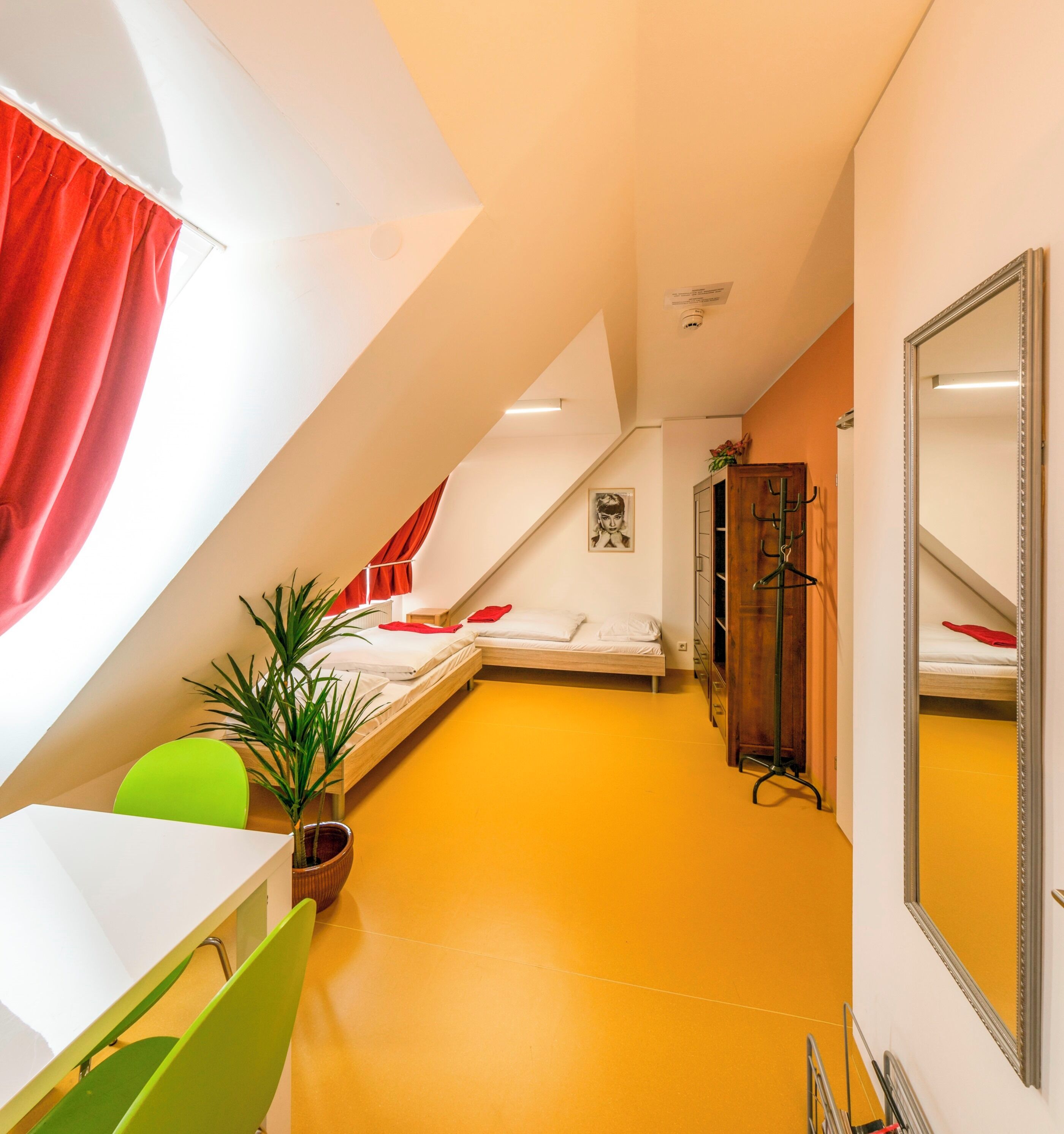 A compact, brightly colored room with two beds, a mirror, a green chair, and a potted plant.