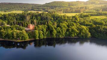 A large, historic building surrounded by dense forest and a calm lake.
