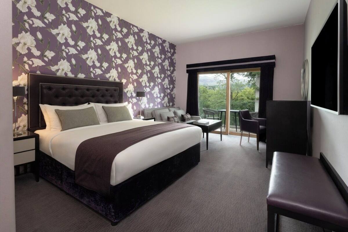 A modern hotel room with a large bed, a desk, a chair, and a balcony with a view of the outdoors.