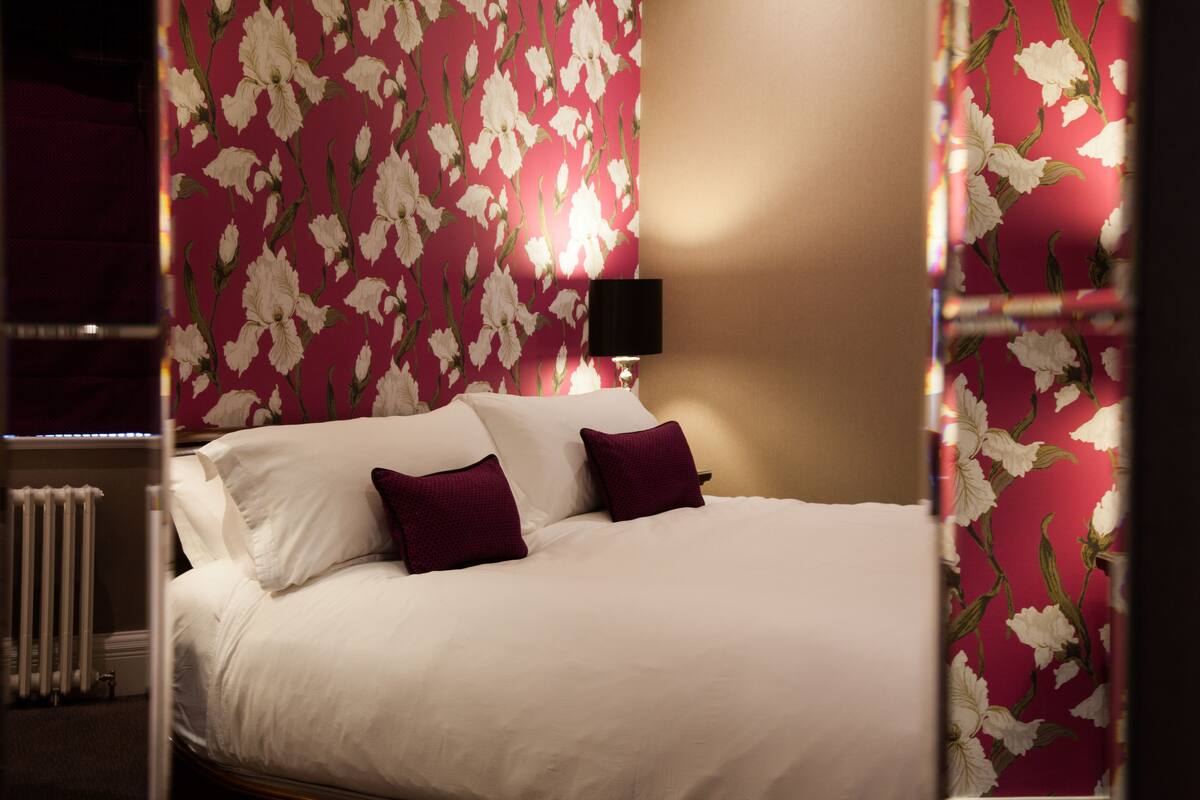 A neatly made bed with white and purple pillows, a black lamp, and a floral wallpaper pattern.