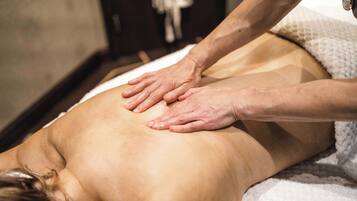 En person får en massage på ryggen medan hen ligger på ett massagebord i ett rum med trägolv och en vit handduk.