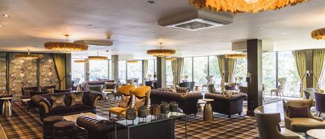 A spacious lounge area with multiple seating options, including sofas and armchairs, arranged around a central coffee table. Large windows provide natural light and views of the outdoors.