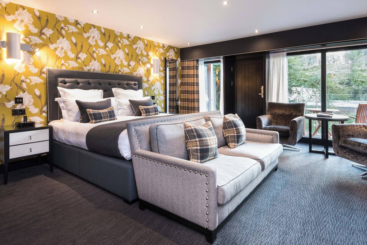 A modern hotel room with a large bed, a sofa, and a chair. There are two windows with curtains, a small table, and a nightstand with a lamp.