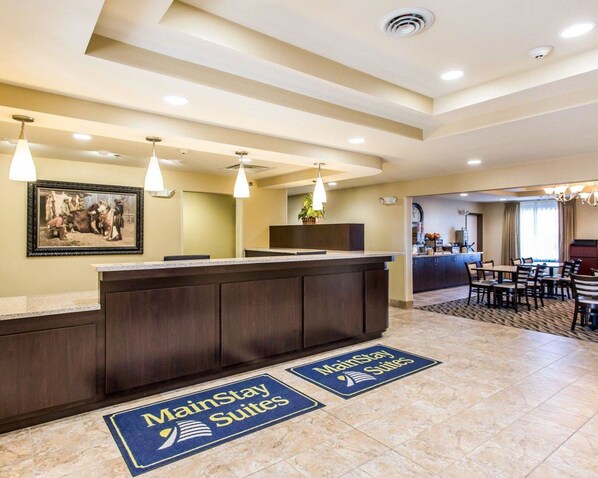 A hotel reception area with a dark wooden reception desk, a framed historical painting on the wall, and a dining area with tables and chairs. - MainStay Suites Rapid City (Rapid City)