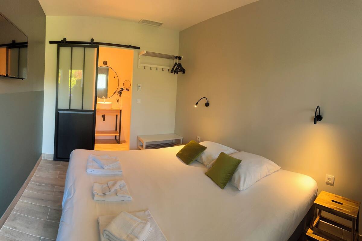 Ein Hotelzimmer mit einem großen Bett, einem Nachttisch, einem kleinen zusätzlichen Nachttisch, einem Spiegel, einem Handtuchhalter, einem Waschbecken und einer Duschkabine.