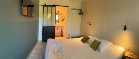 A hotel room with a large bed, a bedside table, a small bedside table, a mirror, a towel rack, a sink, and a shower area.