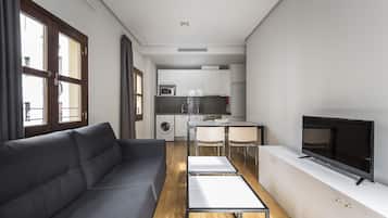 A modern apartment with a grey sofa, a dining area with a table and chairs, a kitchen with white cabinets, and a TV on a white cabinet.