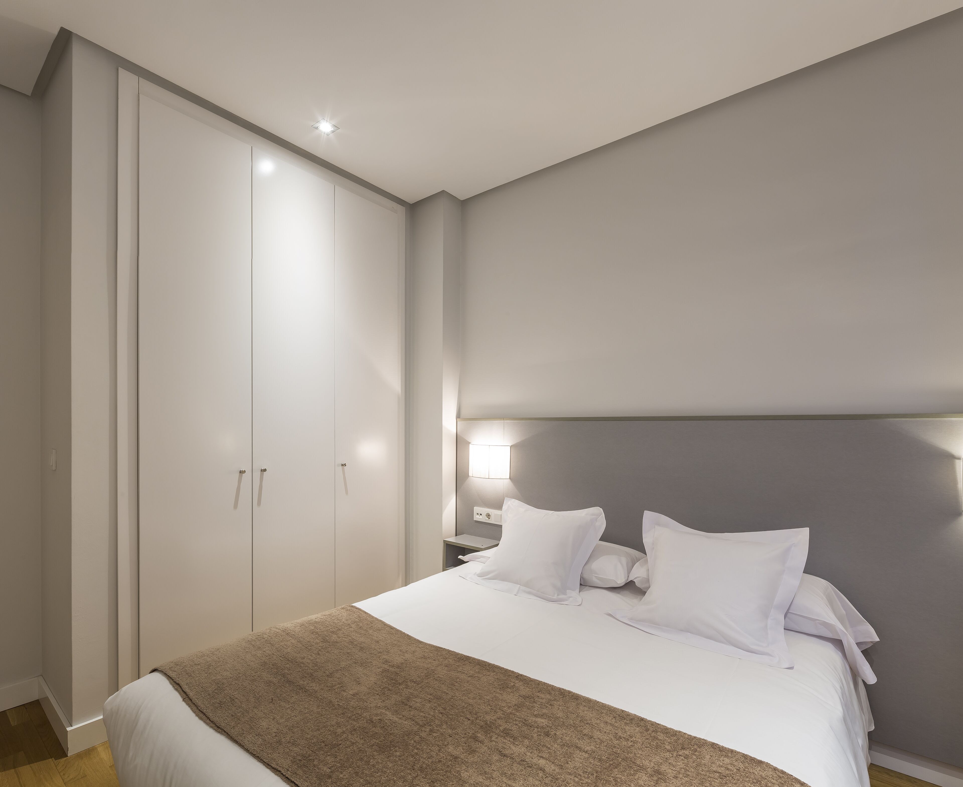 A bedroom with a bed, white bedding, a brown throw blanket, a nightstand with a lamp, and a wardrobe.