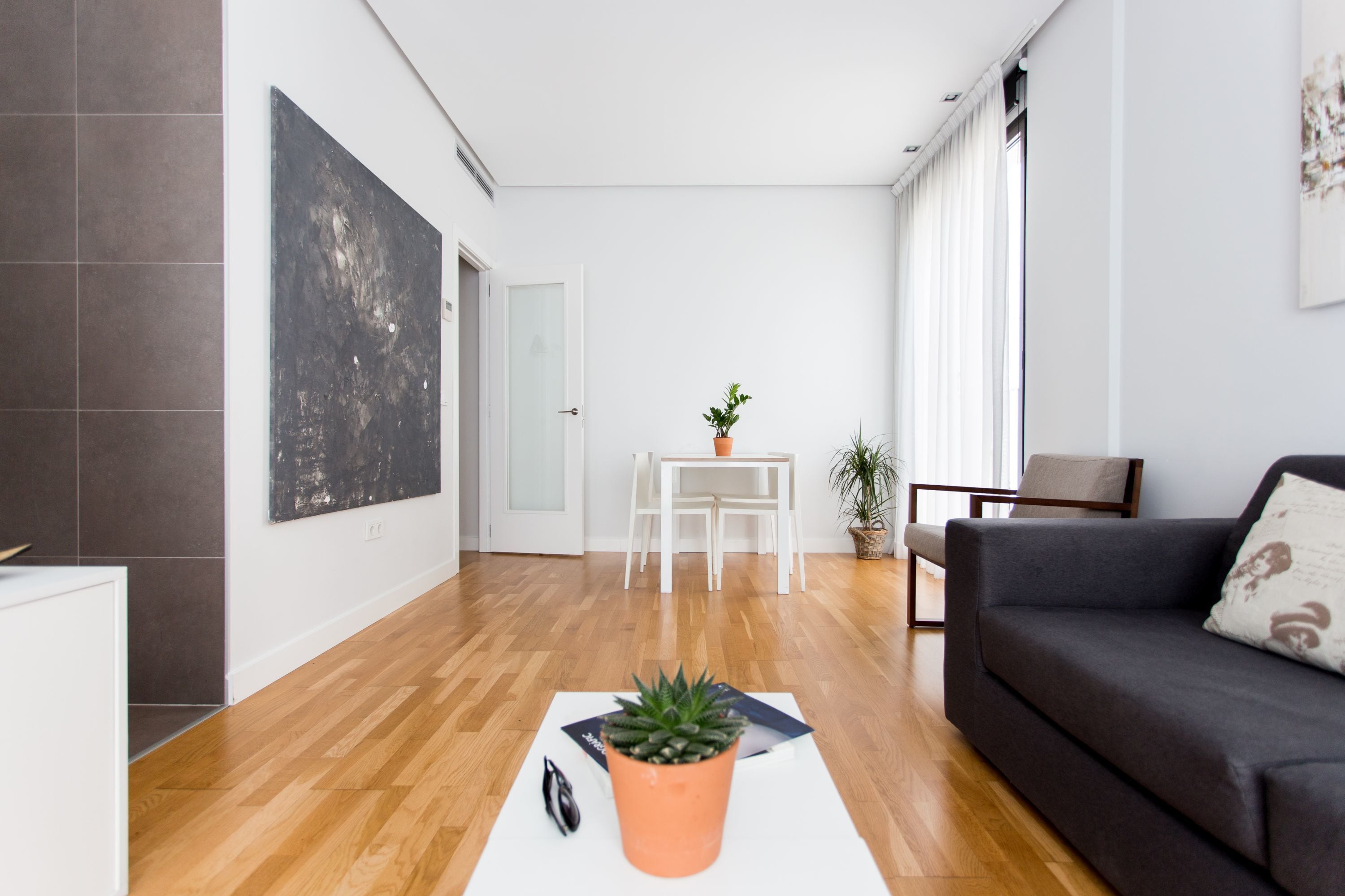Una sala de estar moderna con un sofá oscuro, una mesa de centro blanca con una planta, y un cuadro abstracto grande en la pared.