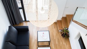 Un salon moderne avec un canapé noir, une petite table ornée d’un objet décoratif, un luminaire suspendu et une vue sur le balcon extérieur.