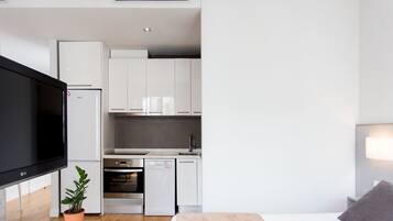 A modern hotel room with a flat-screen TV, a potted plant, a bed with a pink blanket, a small kitchen with white cabinets, and a built-in oven.