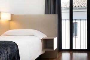 A modern hotel room with a bed, a bedside table, and a view of the outside through a sliding door.