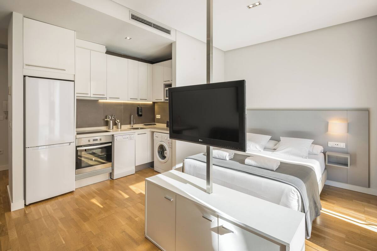 A modern hotel room with a kitchenette, a bed with white bedding, and a flat-screen TV mounted on a cabinet.