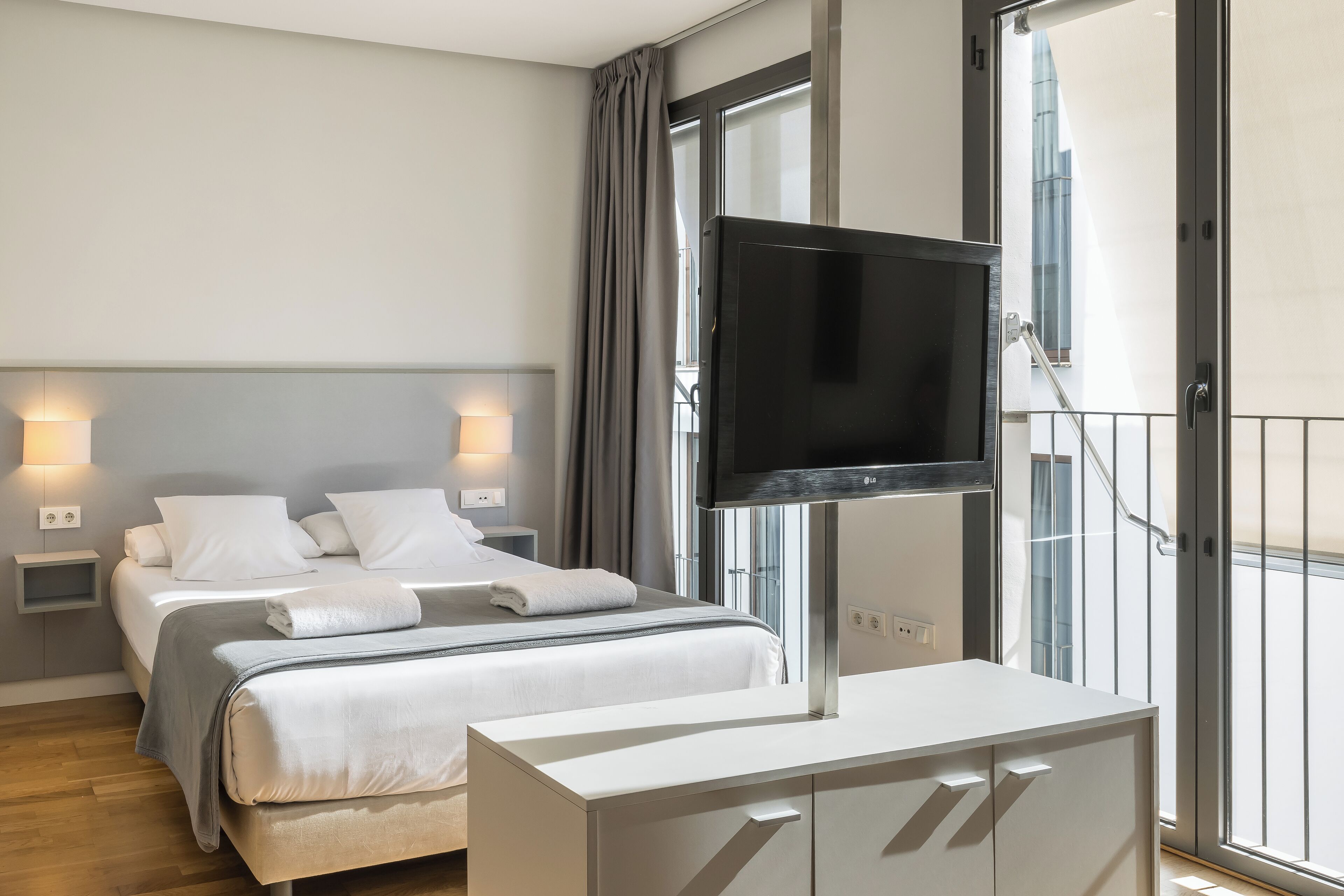 Moderni hotellihuone, jossa on kaksi sänkyä, taulutelevisio, yöpöytävalaisimet ja suuri ikkuna, josta on näkymä rakennukseen.