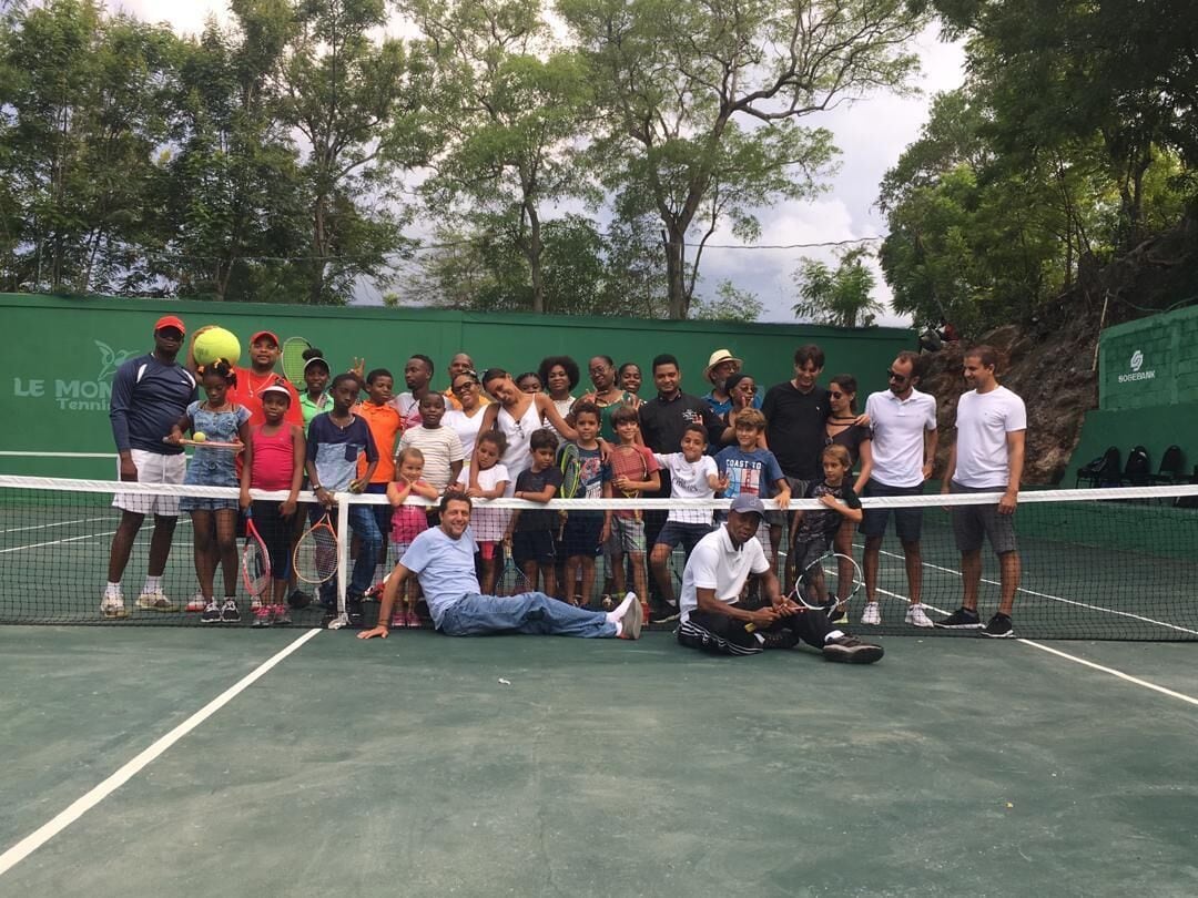 A group of people gathered on a tennis court with a net in the middle, trees in the background, and a green wall with text.
