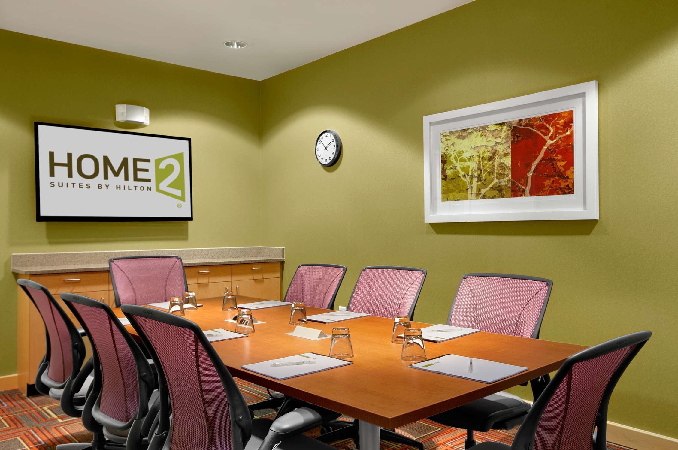 Uma sala de conferência com uma mesa de madeira, seis cadeiras, um relógio, uma obra de arte emoldurada e uma placa com a inscriç ão 'HOME 2 Suites by Hilton'.