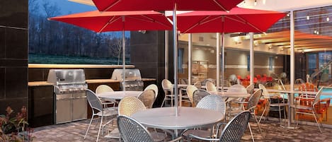 Un patio al aire libre con una parrilla, mesas, sillas y sombrillas rojas.