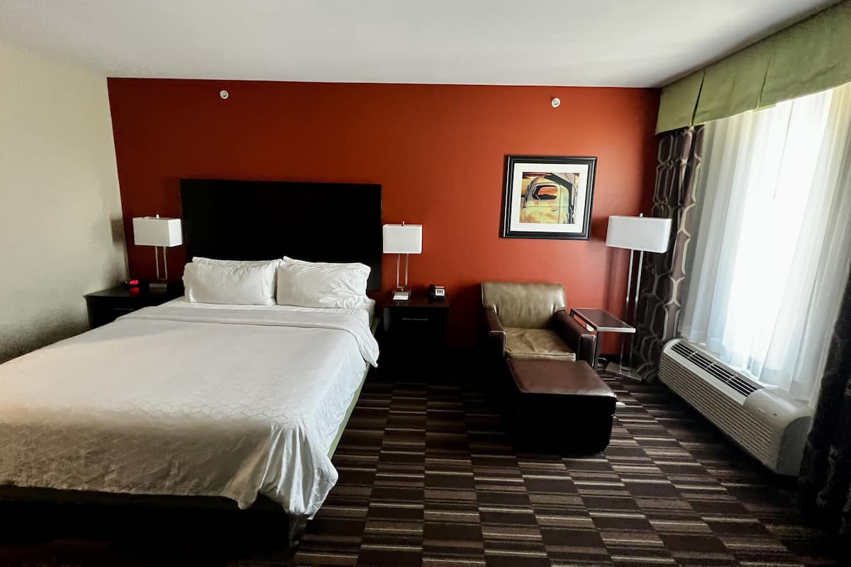 A hotel room with a large bed, two bedside tables with lamps, a chair, a small table, a framed picture on the wall, and a window with curtains.