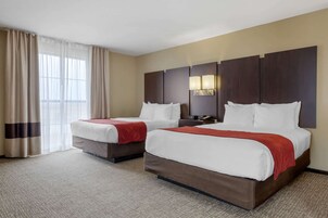 Habitación de hotel con dos camas, paredes color crema, un ventanal con cortinas y una mesita de noche con lámpara.