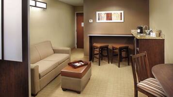 A hotel room with a sofa, ottoman, two chairs, a small table, and a kitchenette area.