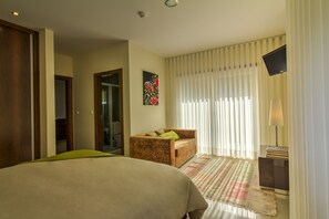 A hotel room with a bed, a sofa, a small table with a lamp, and a window with blinds.