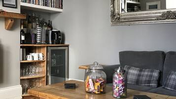 A living room with a wooden coffee table, two glass jars with snacks, a remote control, and a bookshelf with books and bottles.