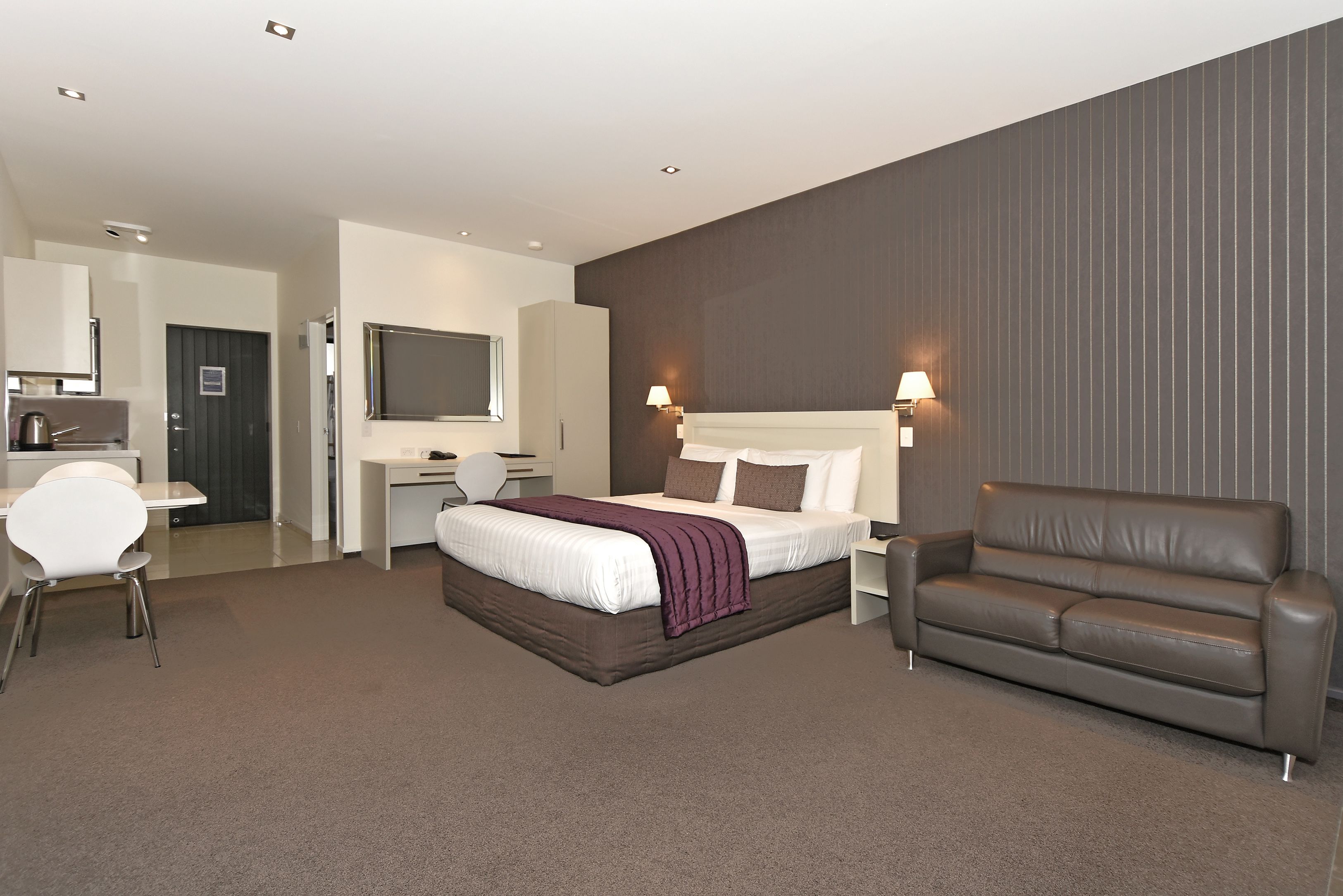 A modern hotel room with a large bed, a brown leather sofa, a desk with a chair, and a TV mounted on the wall.