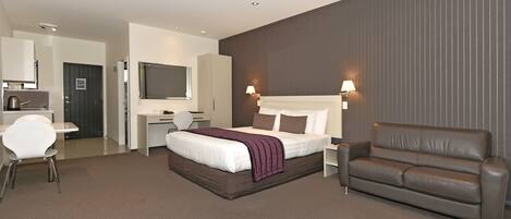 A modern hotel room with a large bed, a brown leather sofa, a desk with a chair, and a TV mounted on the wall.