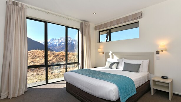 A bedroom with a large window, a bed with a blue throw, and a nightstand with a lamp. - Aoraki Court Motel (Mount Cook)