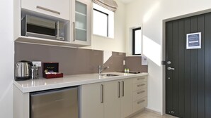 A compact kitchen with a microwave, kettle, and a small refrigerator. There is a sink with a faucet, and a cabinet with drawers. A dark door is visible on the right side. - Aoraki Court Motel (Mount Cook)