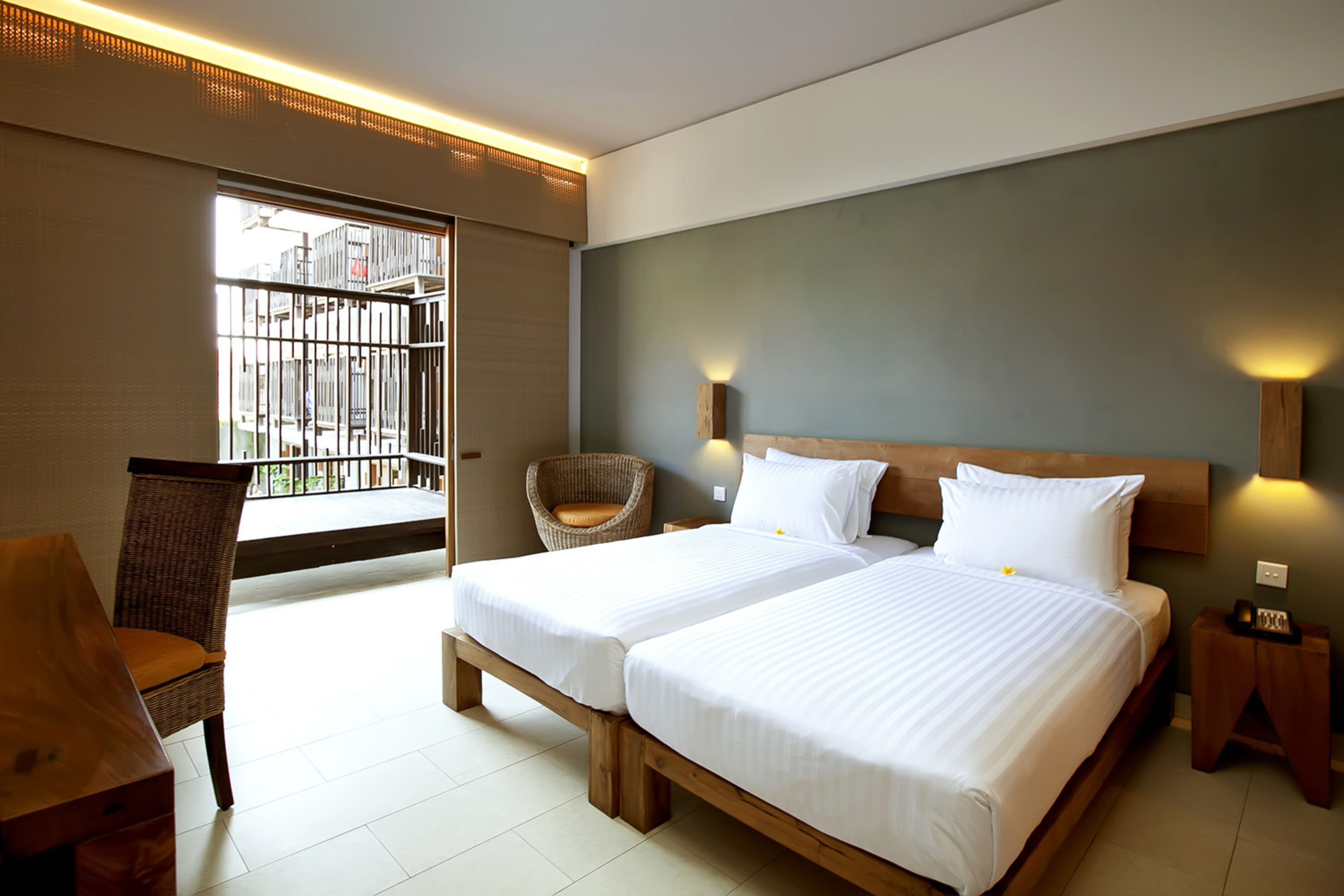 A hotel room with two beds, a wicker chair, a wooden nightstand, and a view of the outside.