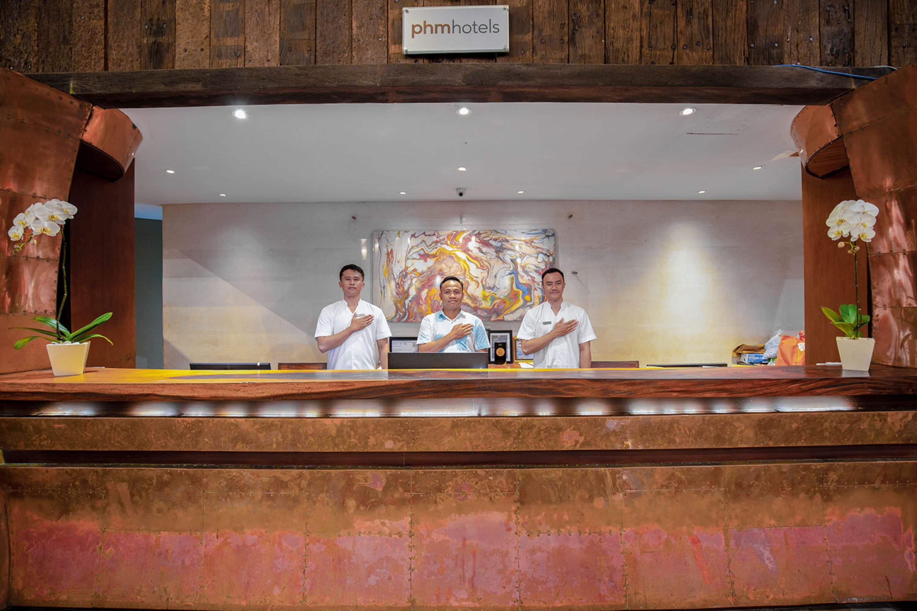 Un mostrador de recepción con tres empleados, un cartel que indica 'phm hotels' y un cuadro abstracto colorido al fondo.