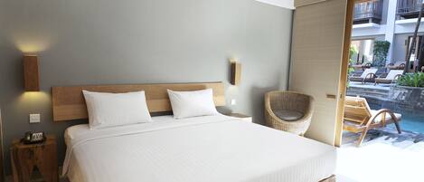 A modern hotel room with a large bed, a nightstand with a phone, a wicker chair, and a view of a pool area.
