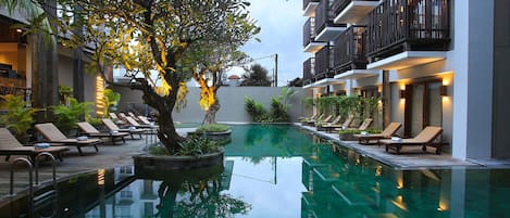 A serene indoor swimming pool with lounge chairs and a tree, surrounded by modern buildings.