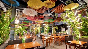 Un comedor interior con mesas y sillas de madera, rodeado de plantas y sombrillas colgantes.