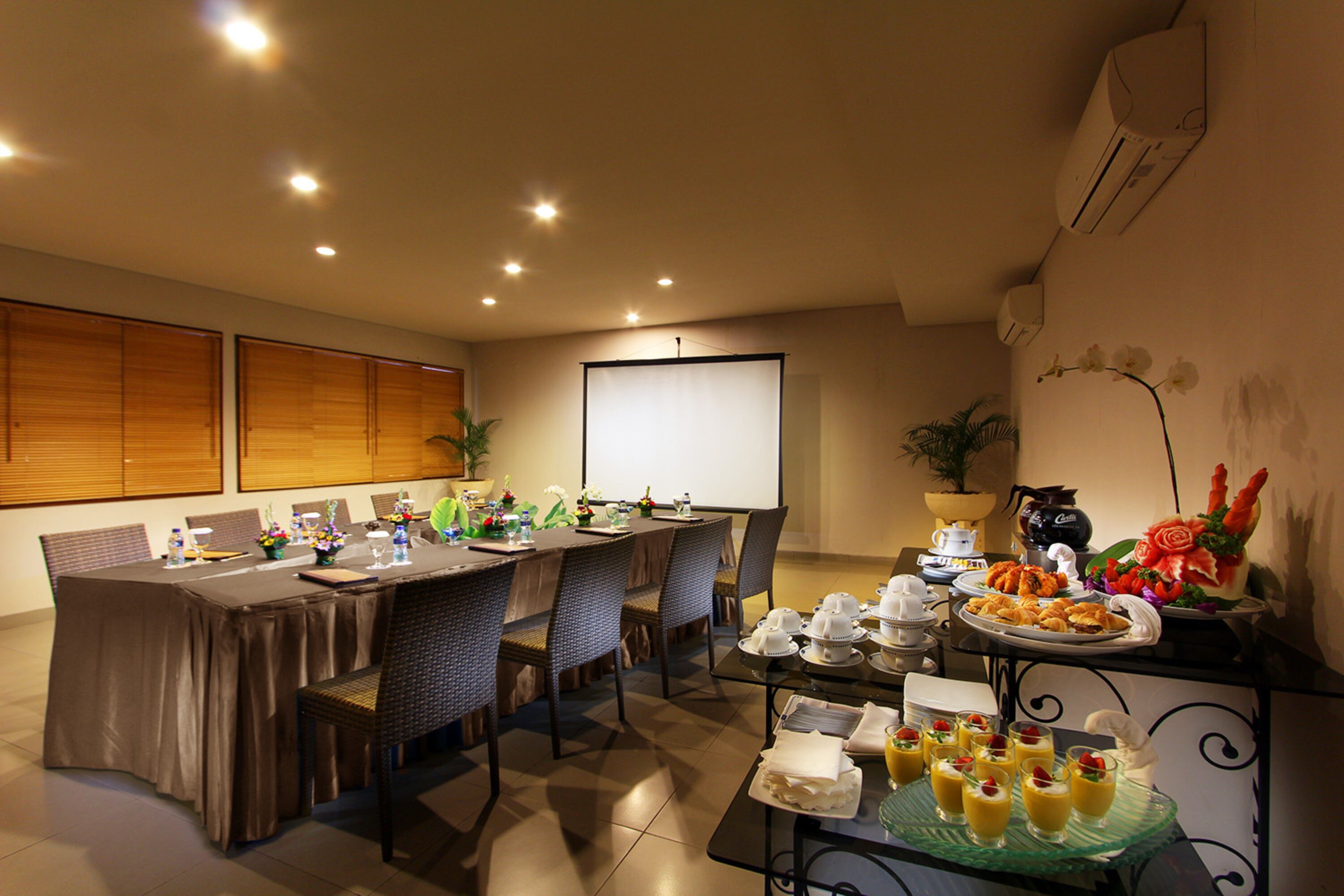 A conference room with a long table, chairs, a projector screen, and a buffet table with food and drinks.