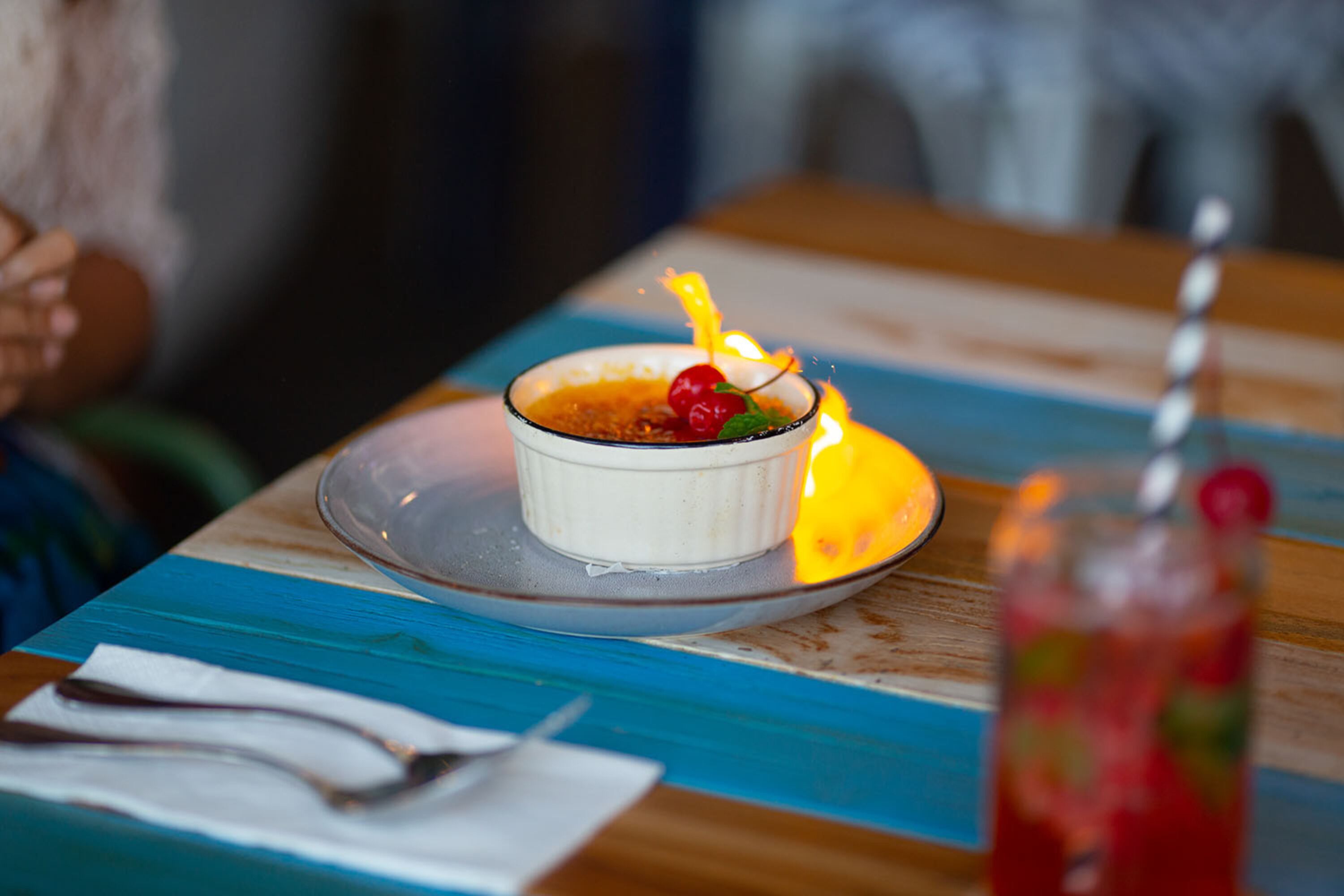 A dessert on a plate with a cherry on top, a spoon, and a glass of red drink on a table.