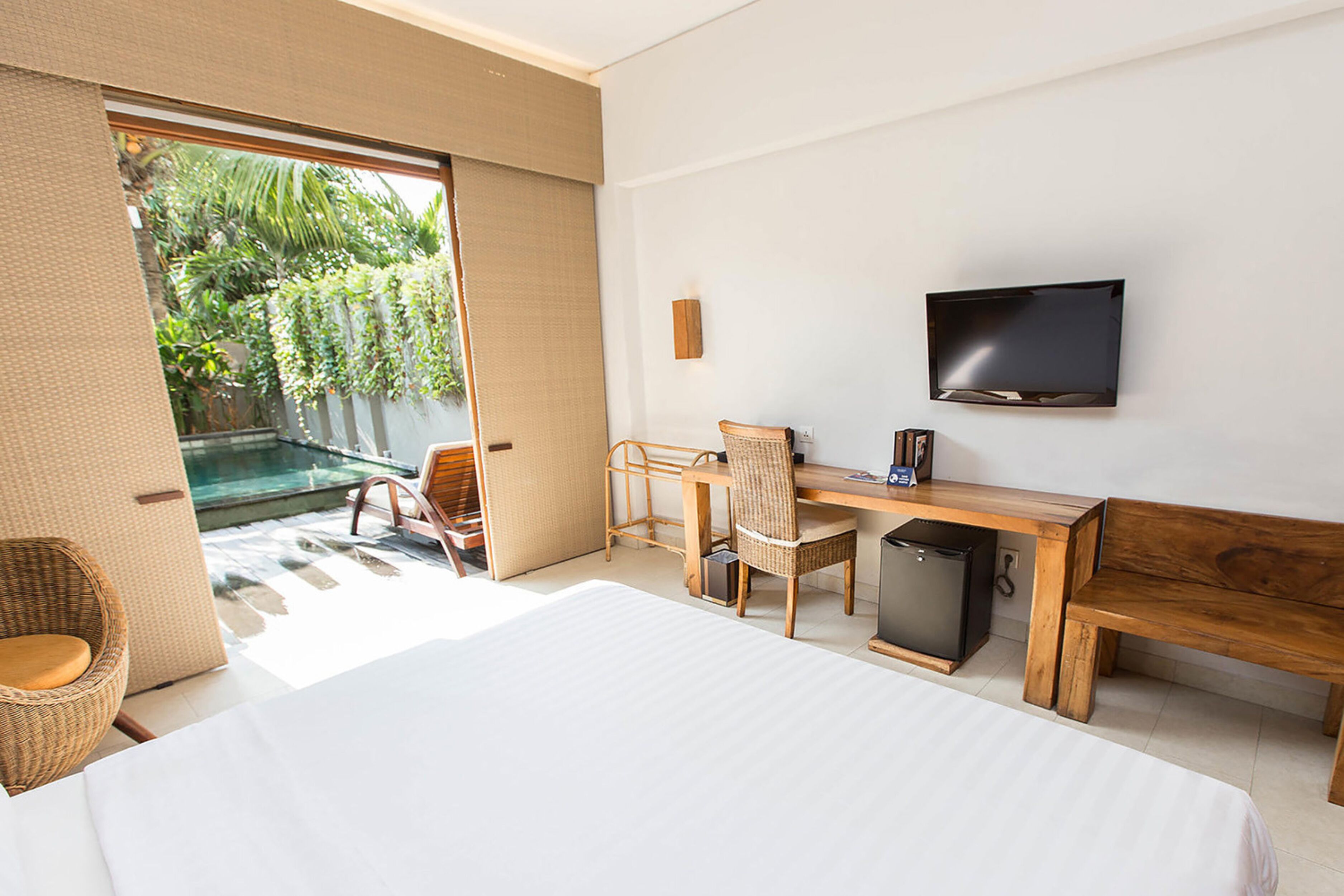 A hotel room with a bed, a desk, a chair, a television, and a view of a pool and greenery outside.