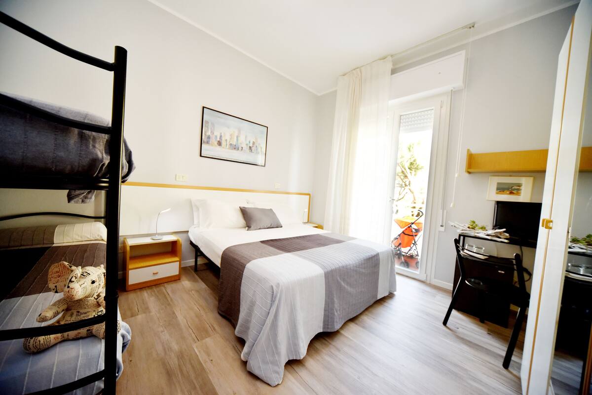 A bedroom with a bunk bed, a single bed, a desk, and a chair. There is a window with curtains and a door leading to a balcony.