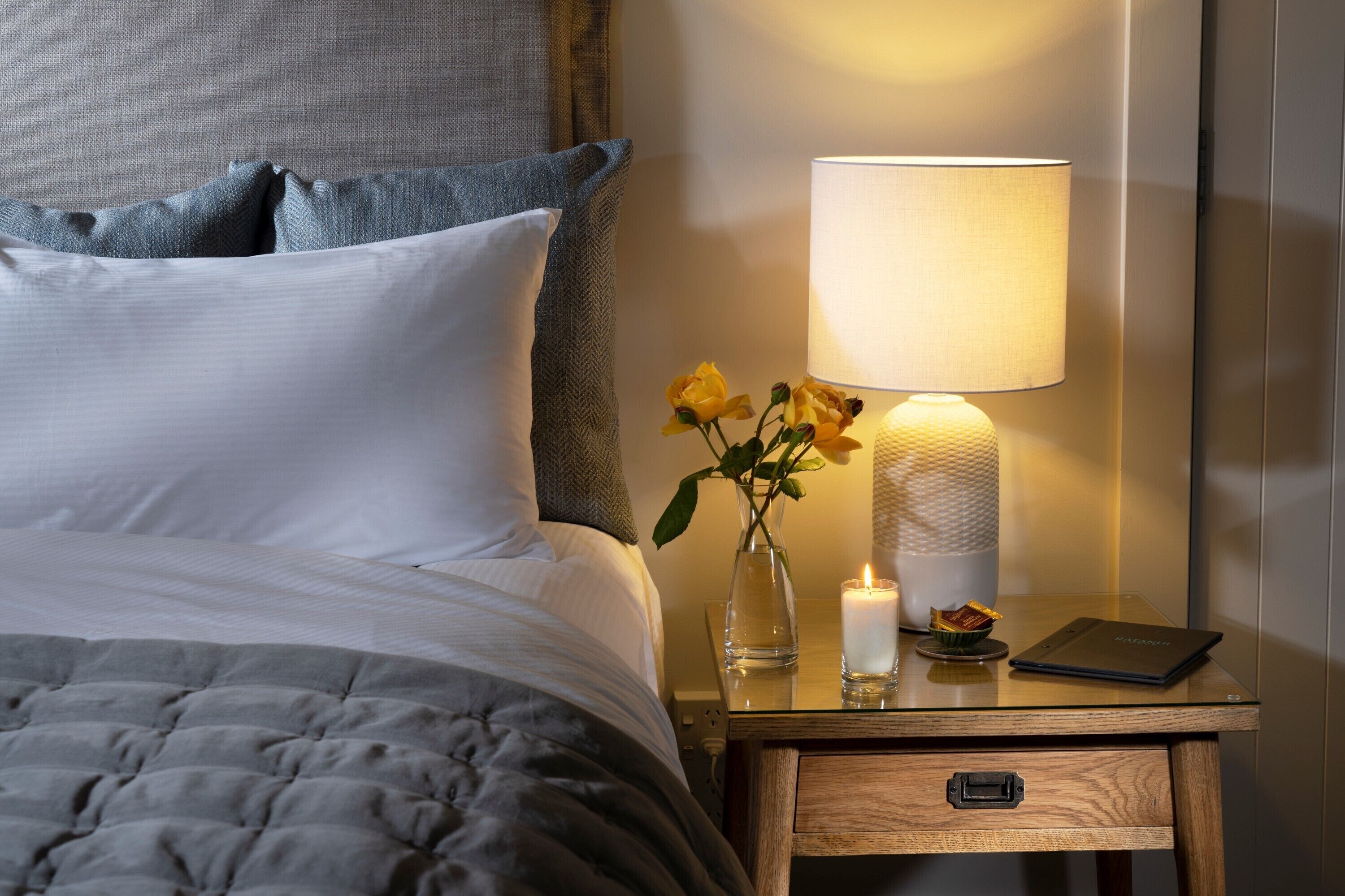 Ein ordentlich bezogenes Bett mit einer grauen Tagesdecke, ein Nachttisch mit Lampe, eine Vase mit Blumen und ein kleines Tablett mit einer Kerze.